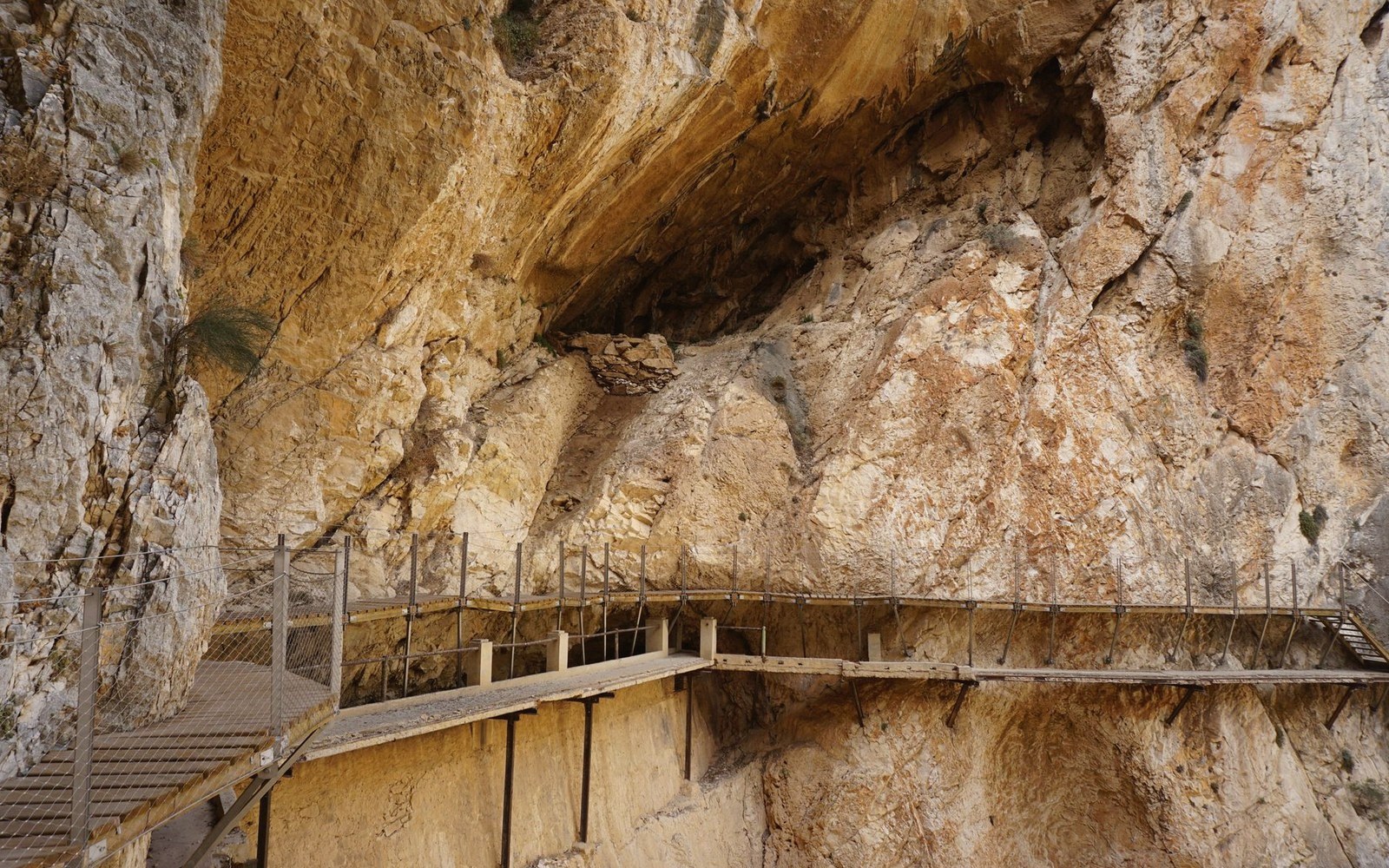A wooden walkway with metal railings clings to a rugged, sunlit limestone cliff face