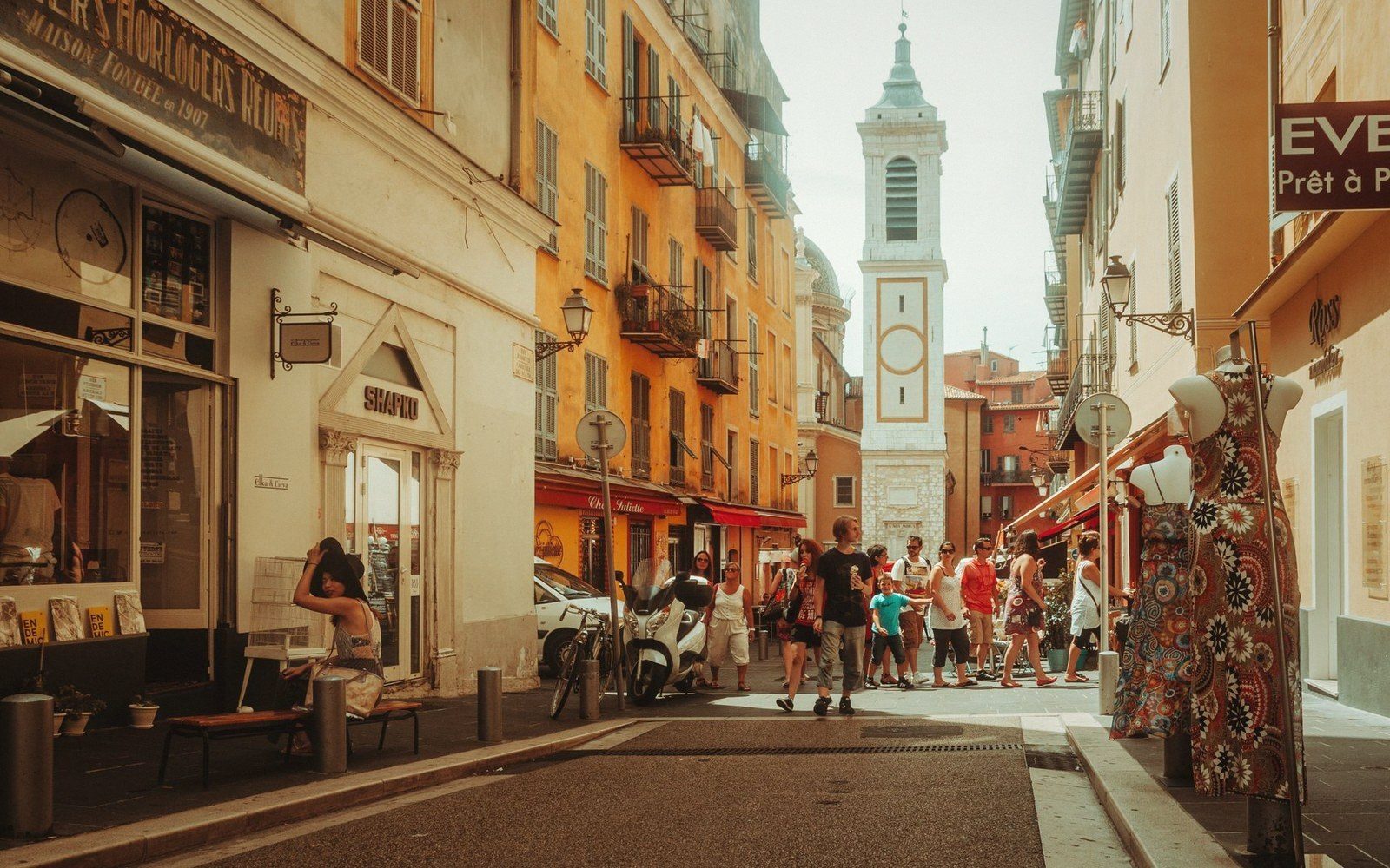 Pedestrians stroll a narrow shopping street lined with ochre buildings and a bell tower