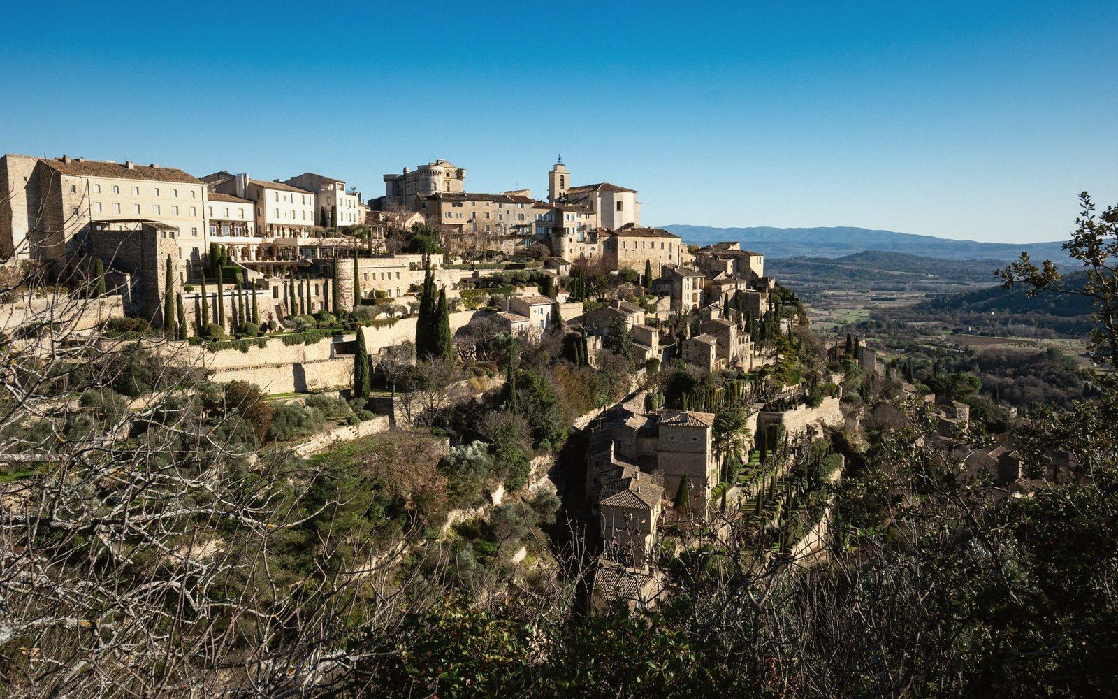 A Luberon perched village seen from across the valley, pale stone against a dark massif ridge, late afternoon